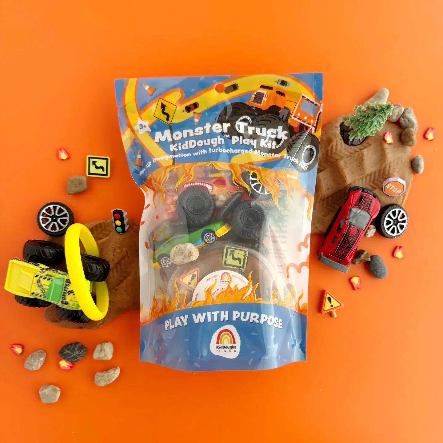The image shows the Monster Truck KidDough Play Kit contents including a bag of textured brown play dough, monster truck, tires, tree, road signs, drive-through ring of fire, jar of fire fimos, crash car, and natural rocks, all arranged on an orange background.