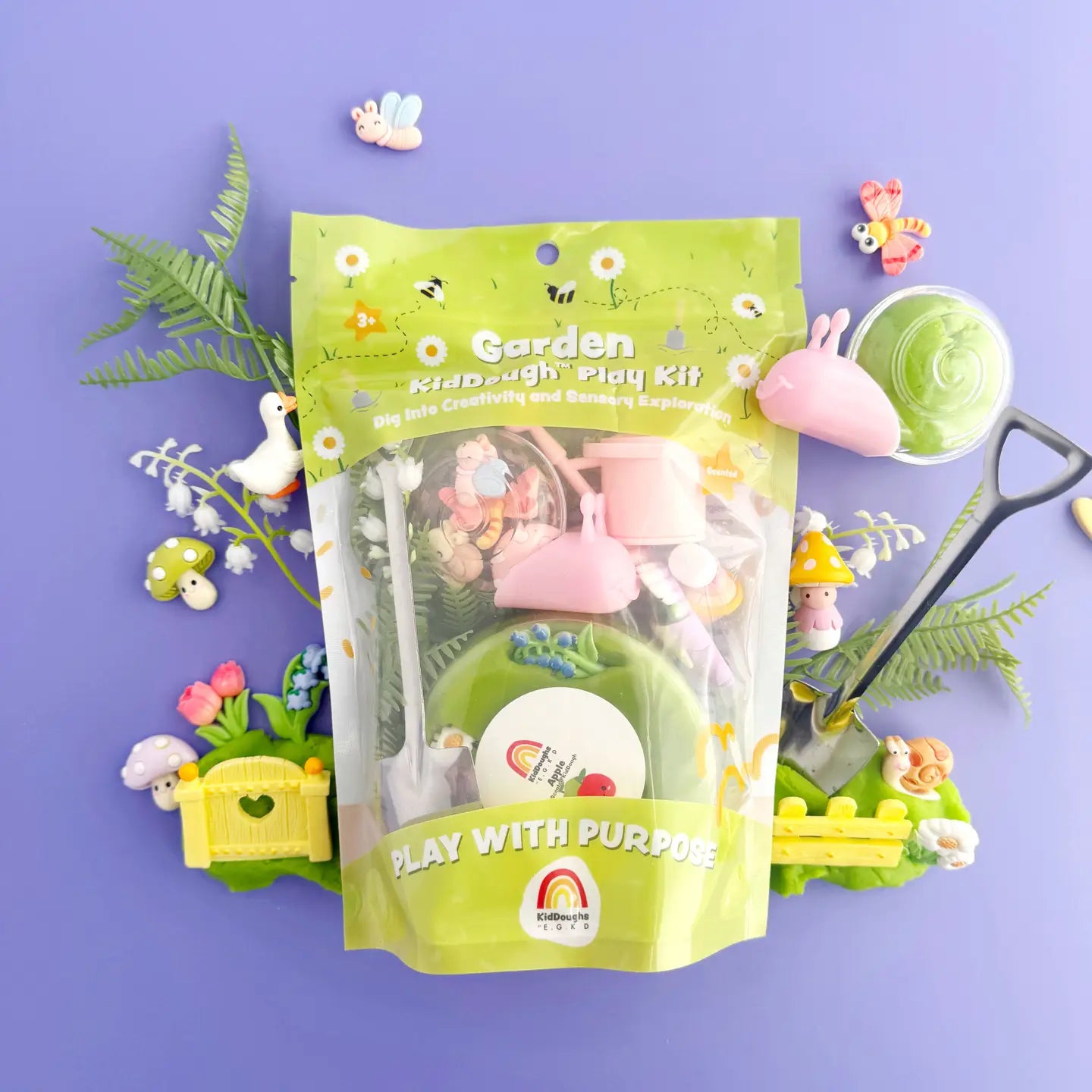 A Garden Kiddough Play Kit with green play dough and various small toy garden items including a garden goose, mushroom, snail charm, and a watering can, displayed on a purple background.