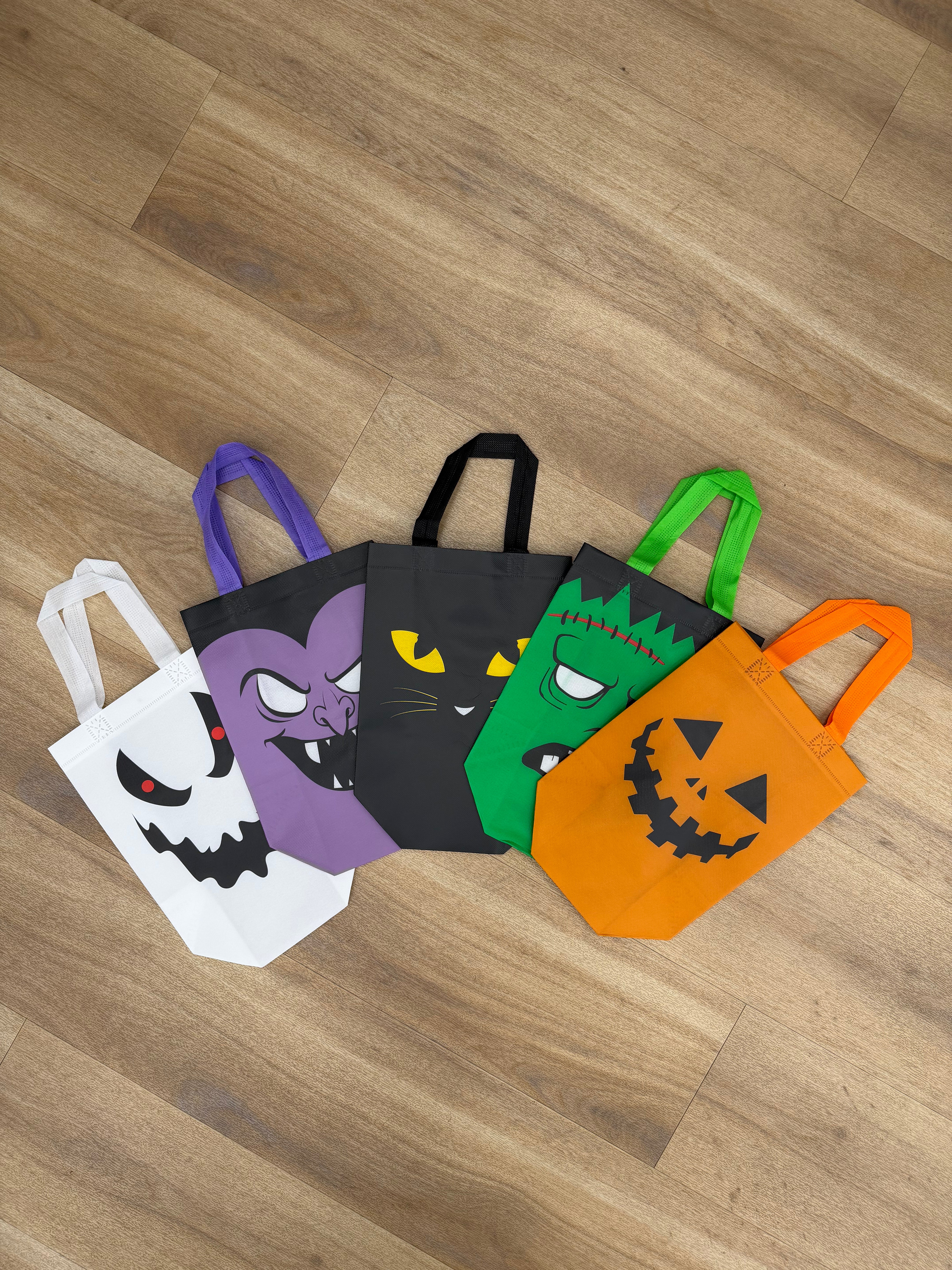 Five Halloween-themed fabric tote bags arranged on a light brown wood floor. The bags are white with a black ghost face, purple with a black cat face, black with yellow eyes, green with a scarface design, and orange with a jack-o'-lantern face.