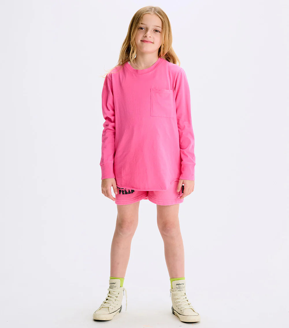 A child in a hot pink long-sleeve t-shirt with a raw, rounded bottom hem and a front pocket, paired with shorts and white sneakers.