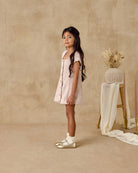 Young girl in a white dress standing against a beige wall with a stool and plant in the background.