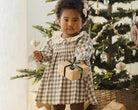 Child in a checkered dress holding a gift box in front of a decorated Christmas tree.