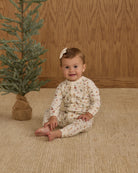 Baby wearing a white holiday romper sitting on a beige rug 