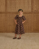 Young girl in a floral dress standing against a wooden paneled wall.