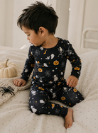 A two-piece children's pajama set featuring a long-sleeve top and matching pants in black with a Halloween print of orange pumpkins, white ghosts, spiders, and moons.