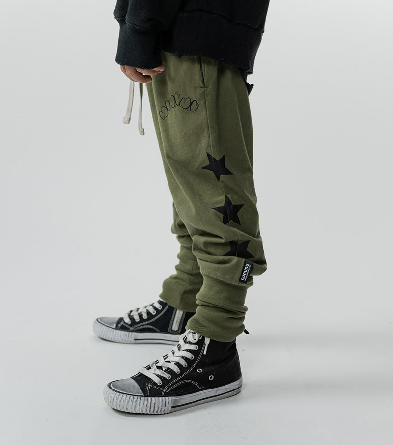 A toddler wearing olive green cotton pants with star patterns on the leg and a black converse sneaker.