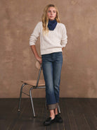 Woman wearing a white sweater and blue jeans standing next to a chair against a brown wall.