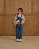 Child wearing blue overalls holding a yellow and brown object against a wooden paneled wall.
