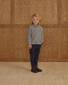 Child wearing a checkered shirt and dark pants standing against a wooden paneled wall.