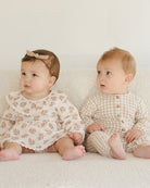 Two babies sitting on a white surface, one wearing a floral dress and the other in a checkered outfit.