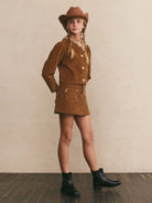 Young girl in a brown outfit with a hat standing against a beige wall.