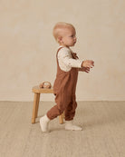 Child wearing brown overalls standing on a wooden stool against a beige background