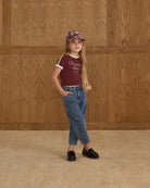 Young girl wearing a burgundy t-shirt with text and blue jeans against a wooden paneled wall.