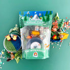 The image shows a Camping KidDough Play Kit which includes various items such as green and blue dough, a bear figurine, a tent, a fire, a stick, foliage, trees, a bear chair, a lantern, s'mores, and trail mix, all displayed on a green background.