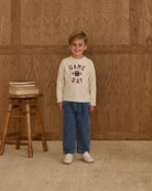 Child wearing a 'Game Day' shirt standing in front of a wooden wall with a stool and books.
