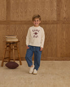 Child wearing a 'Game Day' sweatshirt standing in front of a wooden wall with a stool and football.