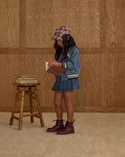 Person in denim outfit with maroon boots standing next to a wooden stool against a wooden paneled wall.