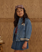 Young girl wearing a denim jacket and skirt with a cap against a wooden paneled wall.