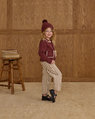 Young girl in a burgundy sweater and striped pants standing against a wooden paneled wall.