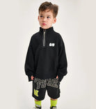 A child wearing a black half zip sweatshirt with a rounded collar, thumbholes, and graphic details on the lower part. The sweatshirt is in a gender-neutral cut.