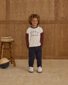 Child wearing a 'Rookie of the Year' shirt standing in front of a wooden paneled wall.