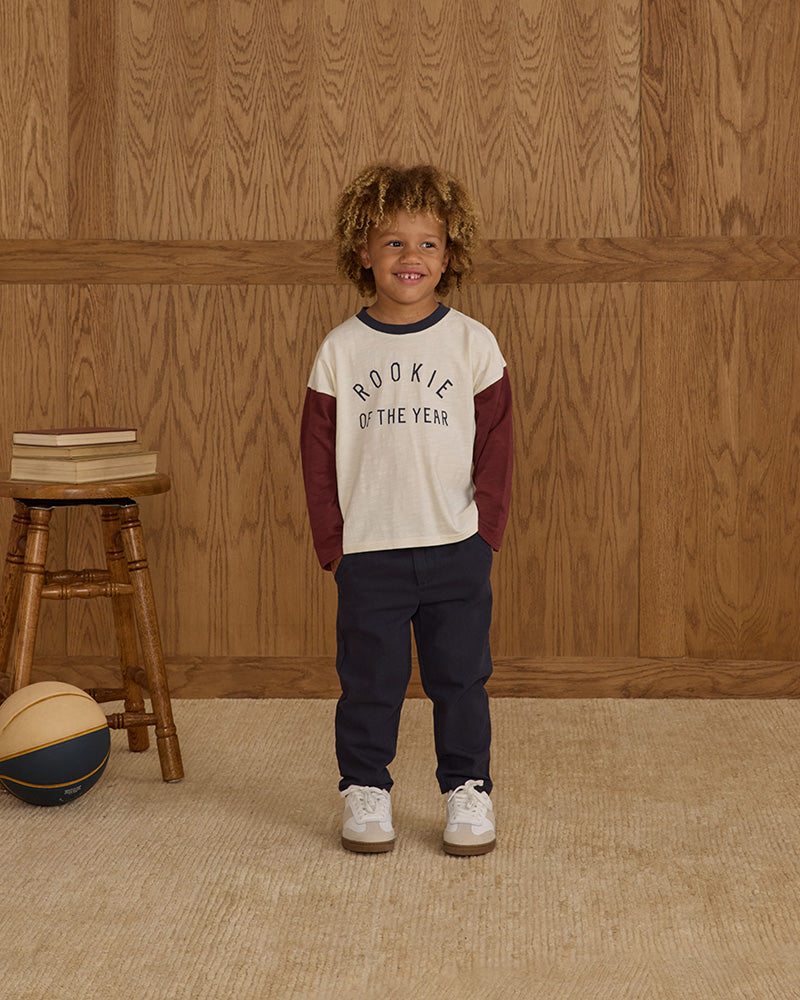 Child wearing a 'Rookie of the Year' shirt standing in front of a wooden paneled wall.