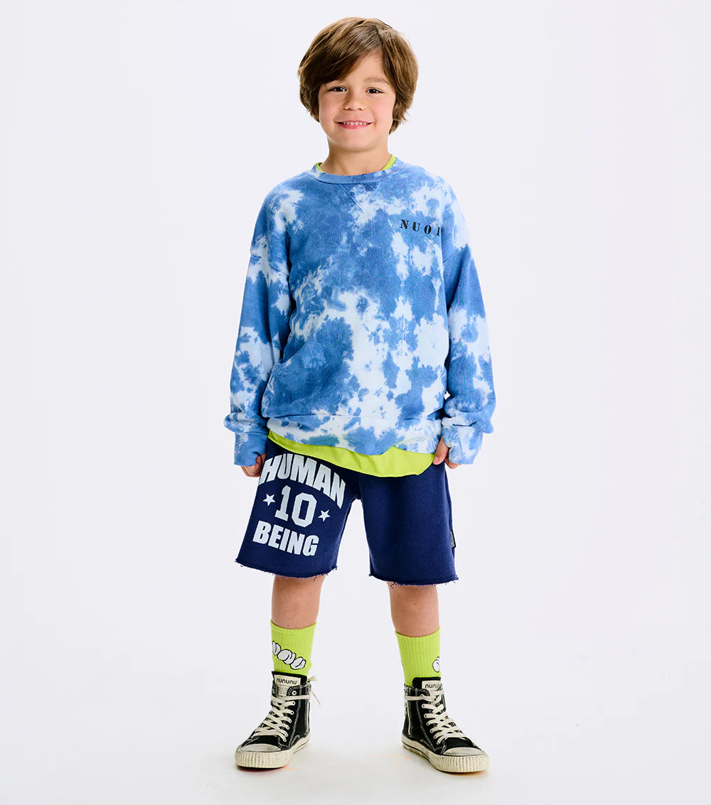 A child wearing a blue tie-dye sweatshirt with 'NU01' printed on the chest, paired with navy shorts, yellow socks, and black sneakers.