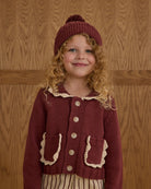 Young girl wearing a maroon knitted cardigan and hat against a wooden paneled wall.
