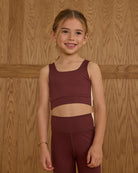 Young girl wearing a maroon athletic outfit standing against a wooden paneled wall.