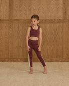 Young girl in maroon athletic outfit standing against a wooden paneled wall.