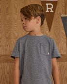 Young boy wearing a navy t-shirt with a small logo, standing against a wooden wall.