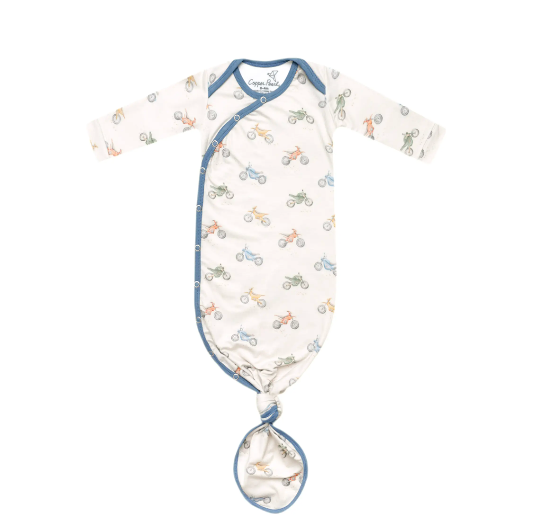 A newborn knotted gown with a pattern of vehicles printed in shades of blue and beige on a white background, featuring a blue binding at the bottom and fold-over hand mitts.