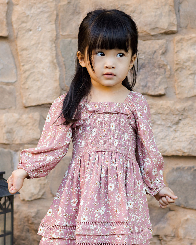 Young girl in a pink floral dress standing against a stone wall.