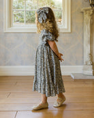 Young girl in a floral dress standing in a room with wooden floor and window.