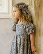 Young girl in a floral dress standing against a stone wall.