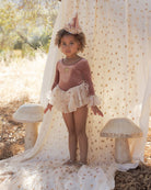 A young girl wearing a dusty rose long-sleeve tutu dress with white lace cuffs, a white ruffled skirt, and a small party hat.