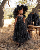 A young girl stands outdoors in a wooded setting wearing a black off-the-shoulder tulle dress covered in silver star-like embellishments, with a large matching black bow headpiece. 