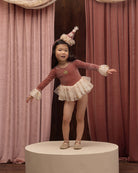 A young girl wearing a dusty rose long-sleeve tutu dress with white lace cuffs, a white ruffled skirt, and a small party hat.