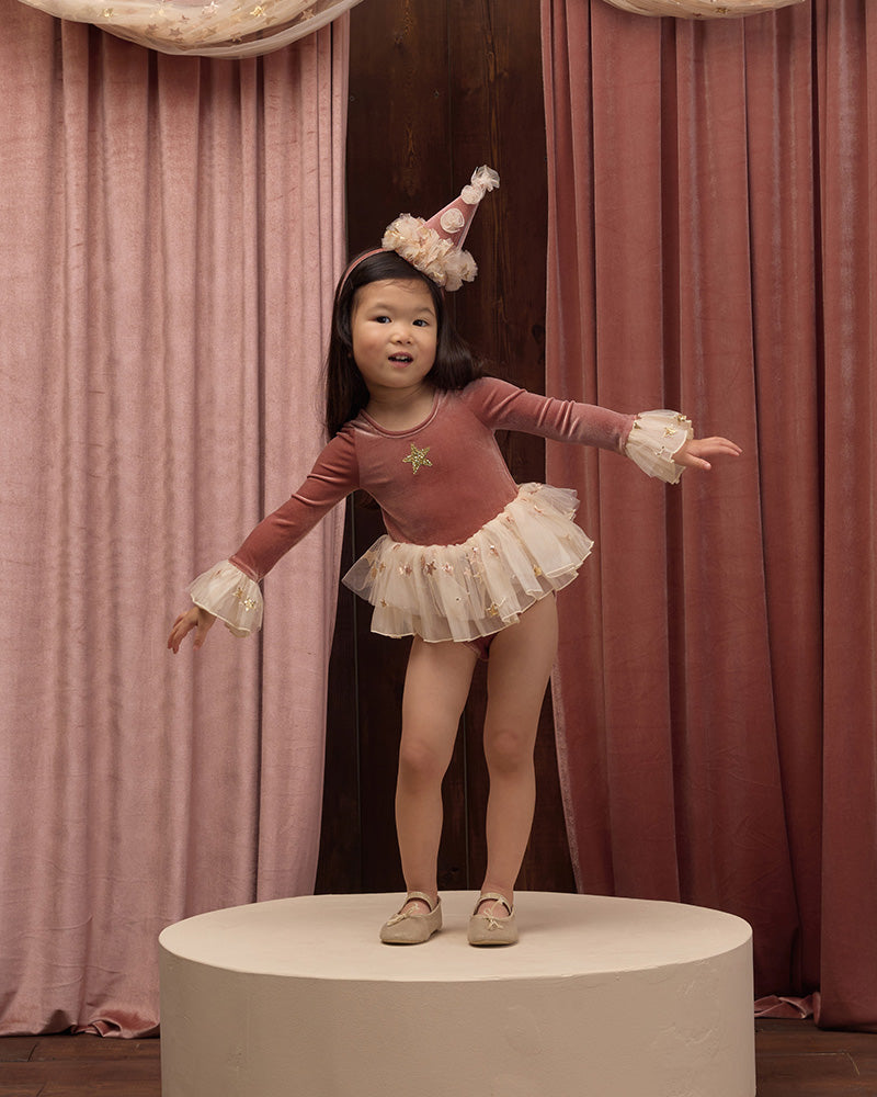 A young girl wearing a dusty rose long-sleeve tutu dress with white lace cuffs, a white ruffled skirt, and a small party hat.