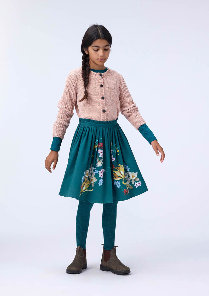 Young girl wearing a pink cardigan, teal floral skirt, and green leggings on a white background