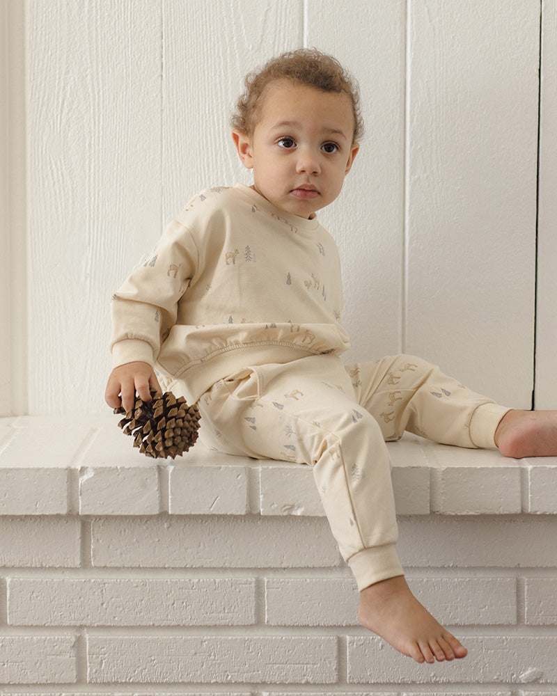 Off-white jogger pants for toddlers, featuring an all-over print of small deer and pine trees in muted gray and beige tones, with an elastic waistband and drawstring.