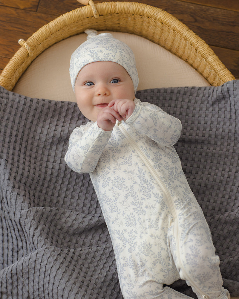 A baby wearing a light cream zip-up footed sleeper and matching hat with a delicate light blue floral pattern, lying on a textured gray blanket inside a woven basket.