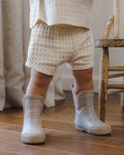 A pair of children's rain boots featuring a light gray and beige plaid pattern with solid beige elastic side panels and soles, shown on a plain white background.