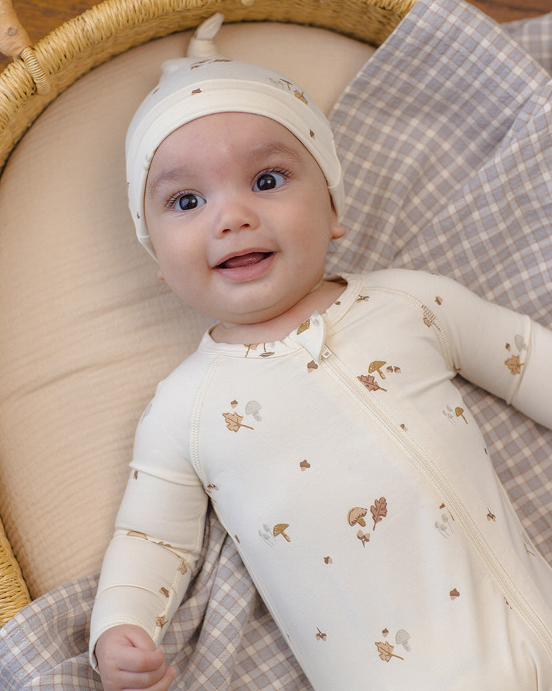 A baby wearing a cream-colored zip-up bodysuit and matching hat with small brown woodland prints, set against a beige and gray check blanket.
