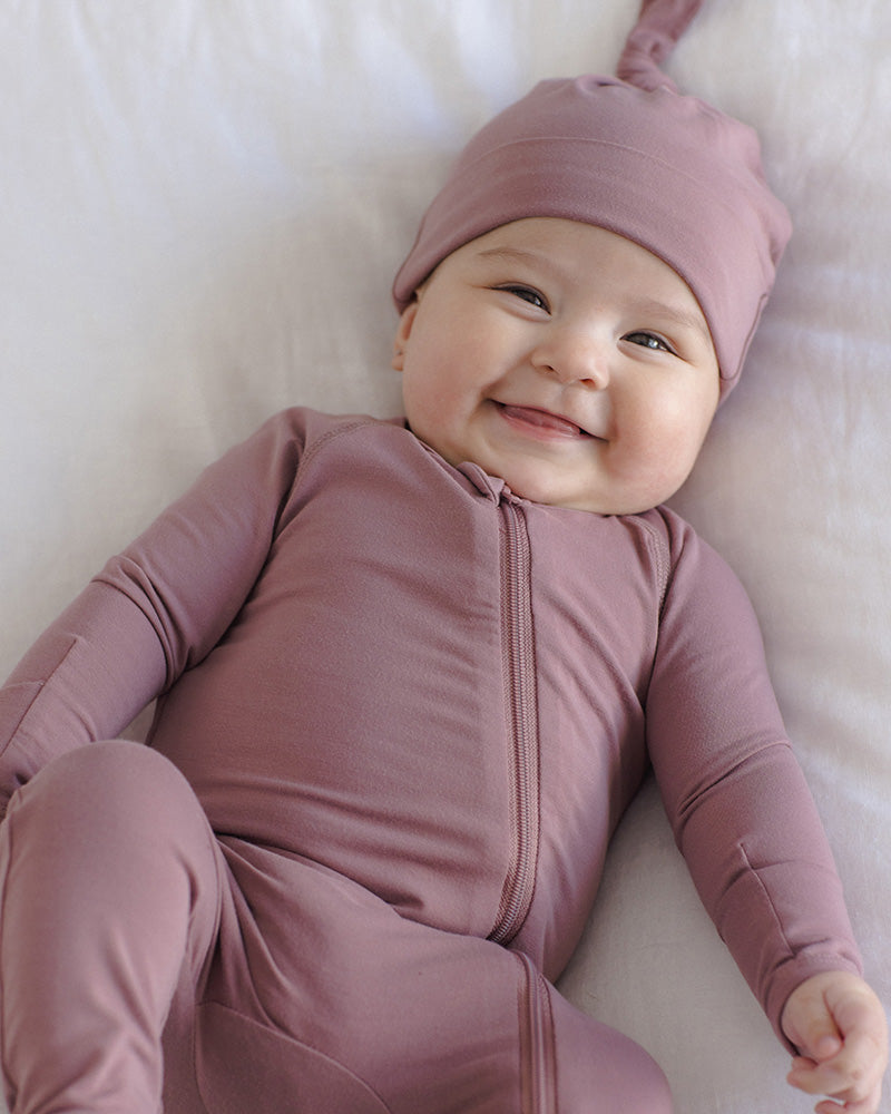 Baby lying on a white blanket, wearing a dusty mauve zip-front footed onesie and matching pointed hat.