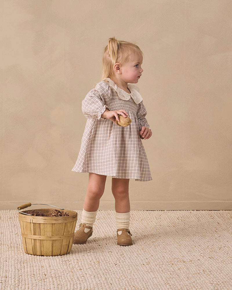 A young blonde girl seen from behind wearing a beige and white plaid dress with a large white ruffled collar, set against a solid beige background.