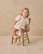 A baby girl's short-sleeve dress in cream cotton fabric with a delicate gray floral print, featuring a smocked bodice and puffed sleeves.