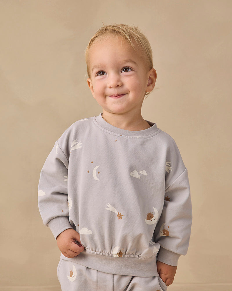 A light gray long-sleeve sweatshirt for toddlers, featuring an all-over celestial print of white clouds, crescent moons, and brown stars.