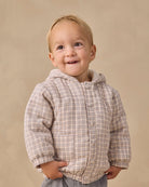 A smiling toddler wearing a beige and brown plaid hooded button-up jacket, standing against a neutral beige studio background.