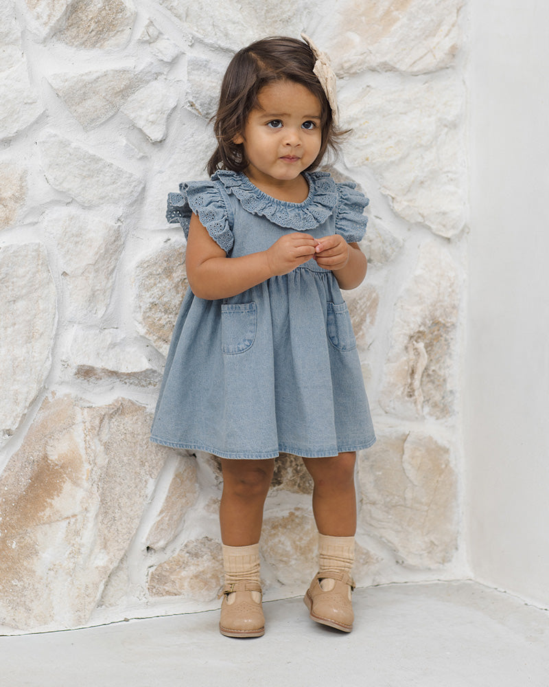 Young girl wearing a light blue dress with ruffled collar against a stone wall.